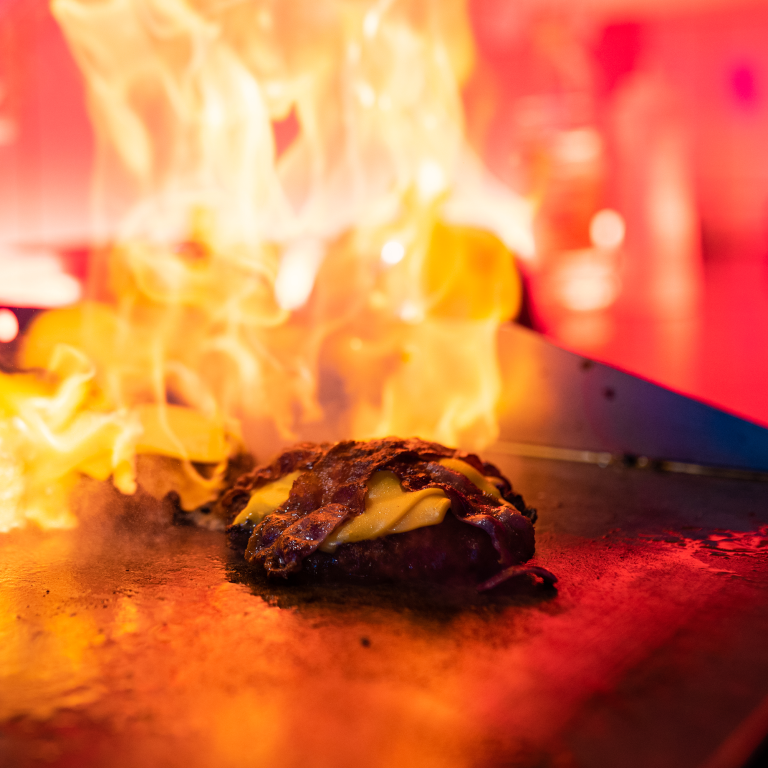 Ein saftiger Burger-Patty auf einem Grill mit Flammen, fotografiert während eines Food-Fotoshootings für Tommy's Braterei. Hochauflösende Fotografie für Social Media von JNPhotografics.