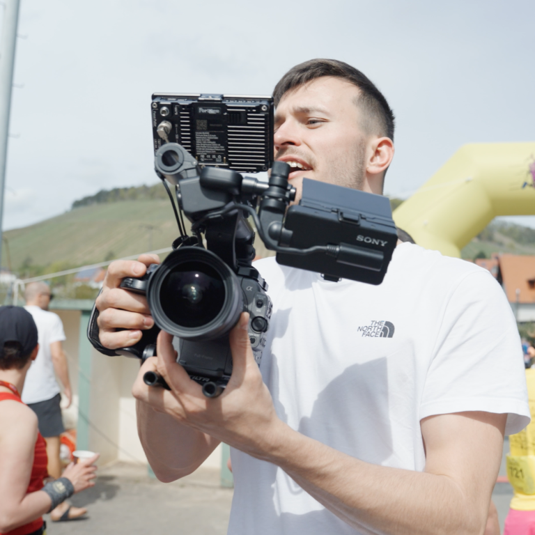 Ein Bild von Johannes Nöth, der eine Filmkamera bei einem Sportevent hält. Im Hintergrund sind verschiedene Aktivitäten und Menschen zu sehen. Der Alt-Text könnte lauten: 'Johannes Nöth filmt hinter den Kulissen für den Sportevent-Aftermovie, während Athleten und Zuschauer im Hintergrund agieren