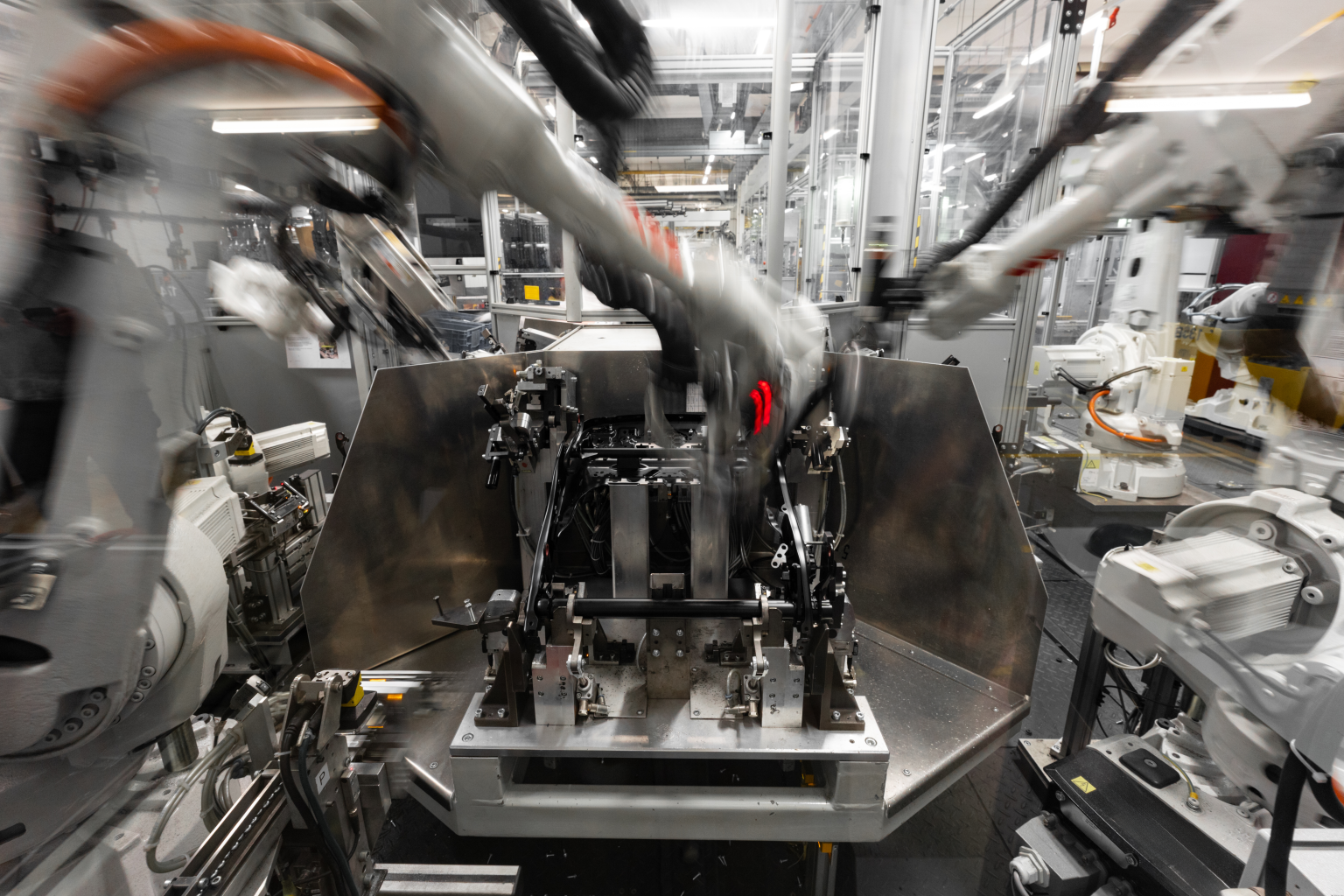 Industrieroboter bei der automatisierten Fertigung eines Fahrzeugbauteils in der Produktionshalle von Brose Automotive in Coburg. Der orangefarbene Roboterarm montiert präzise ein Metallteil an einer industriellen Vorrichtung. Im Hintergrund befinden sich weitere Robotereinheiten und Maschinenkomponenten – ein typisches Beispiel für Industrie 4.0 im Automotive-Bereich. Hochwertige Industriefotografie mit Fokus auf Automatisierung, Präzision und technische Effizienz. Fotografiert von Johannes Nöth, JNPhotografics.de, mit Sony A7 IV und dem Sony GM 70–200mm f/2.8.