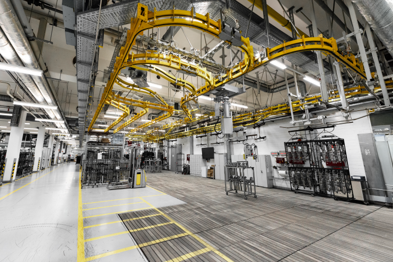 Industrie Fotografie einer Produktionshalle bei Brose Automotive in Coburg: Weitwinkelaufnahme zeigt gelbes Trägergestänge an der Hallendecke, an dem Bauteile hängend durch eine automatische Förderanlage bewegt werden – vermutlich Teil einer Lackieranlage oder Trocknungslinie. Der Blick in die moderne Fertigung vermittelt Tiefe, Struktur und technische Präzision. Diese professionelle Industrie Fotografie von Johannes Nöth, JNPhotografics.de, wurde mit einer Sony A7 IV aufgenommen und zeigt eindrucksvoll industrielle Abläufe in der Automobilbranche. Perfekt geeignet für Unternehmenskommunikation, Imagekampagnen und Webauftritte mit Fokus auf hochwertige Industrie Fotografie.